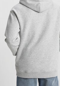 Sudadera gris con un bolsillo canguro y capucha con cordón, hecha de una tela suave, con puños y dobladillo acanalados, vista desde la parte de atrás.