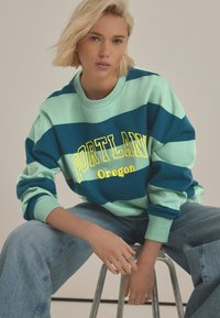 Přehnaně velký svetr v teal a světle zelených pruzích s nápisem "PORTLAND" a "Oregon" vytištěným žlutou barvou. Má žebrované manžety a límec.