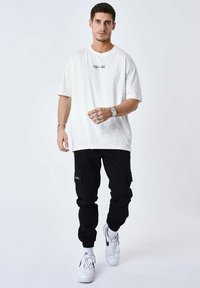 T-shirt bianca in cotone con testo grafico nero, abbinata a pantaloni cargo neri con tasche laterali e sneaker sportive bianche.