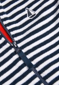 Vêtement zippé rayé bleu marine et blanc avec une fermeture éclair rouge, comportant un détail de bateau à voile brodé sur une texture douce de serviette.