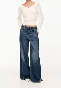 Cream knitted crop sweater with a wide V-neck, paired with dark blue wide-leg jeans featuring front pockets and distressed accents.