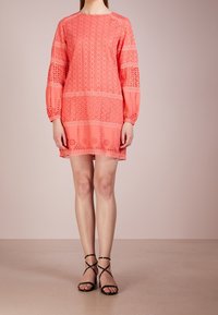 Růžové korálové šaty s dlouhými balónovými rukávy, zdobené výřezovými vzory a vrstveným designem. Kombinováno s černými sandály na tenkých páskách. Hladká textura.