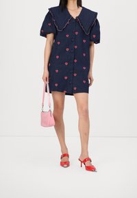 Personne portant une robe bleu marine avec des motifs en forme de cœur rouges, tenant un sac à main rose et des mules à talons hauts rouges avec des embouts métalliques.