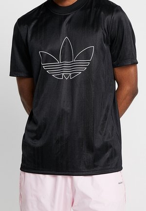 Czarna koszulka z krótkim rękawem z białym logo trefoil Adidas noszona przez osobę z założonymi rękami, w połączeniu z jasnoróżowymi szortami.