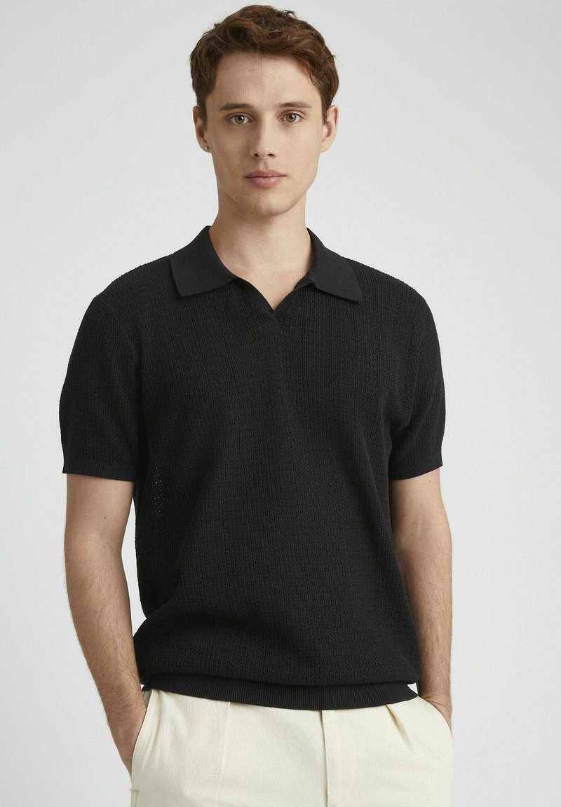 Polo shirt noir à manches courtes avec un design en tricot texturé, présentant un col classique et une coupe décontractée, associé à un pantalon de couleur claire.