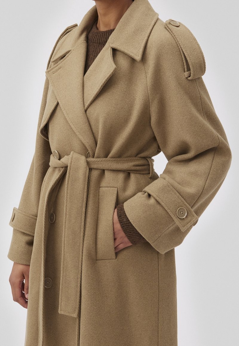 Manteau trench en laine beige avec un design croisé, grands revers, poches latérales et poignets boutonnés. Tissu texturé avec une taille ceinturée.