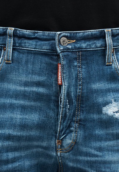 Κοντινή λήψη μπροστινού μέρους από μπλε τζιν distressed με κουμπί, φερμουάρ, θηλιές για ζώνη και κόκκινη ετικέτα με το λογότυπο DSQUARED2.