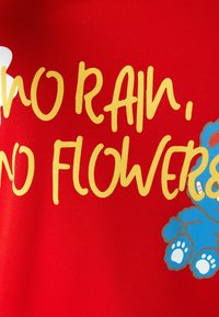 Camiseta de tela roja con el texto amarillo "NO HAY LLUVIA, NO HAY FLORES" y un gráfico de un oso de peluche azul con acentos blancos en sus patas.