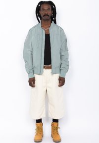 Lichtblauwe bomberjack, zwarte tanktop, witte baggy shorts en gele veterschoenen. Accessoires zijn een zilveren ketting en oorbellen.