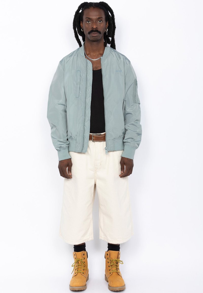 Chaqueta bomber azul claro, top negro sin mangas, pantalones cortos blancos holgados y botas amarillas con cordones. Los accesorios incluyen una cadena de plata y pendientes.