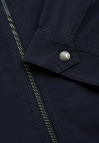 Dark navy fabric jacket detail with metal zipper and flap secured by a silver snap button engraved with brand text.