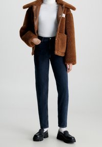 Person klädd i brun fleecejacka, vit polotröja, svarta slim-fit jeans, vita strumpor och svarta plattform loafers på vit bakgrund.