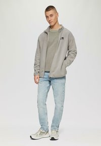 Jasnoszara polarowa kurtka z zamkiem i naszywką z logo, noszona na zielonym swetrze. Jasnoniebieskie dżinsy i beżowe trampki dopełniają stylizację.