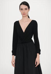 Crna haljina s dubokim V-izrezom, dugim rukavima, detaljem uvrta u struku i lepršavom suknjom. Glatka tekstura tkanine s mat završetkom.