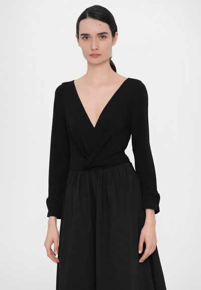 Juoda suknelė su gilia V formos iškirpte, ilgomis rankovėmis, pynės detale ties juosmeniu ir laisvai krentančia sijono dalimi. Lygi audinio faktūra su matinio paviršiaus apdaila.