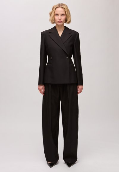 Femme aux cheveux blonds courts portant un blazer noir cintré à double boutonnage et un pantalon noir à jambes larges, debout devant un fond uni.