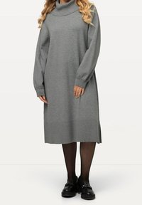 Robe en tricot gris avec un col bénitier, des manches longues, des poignets côtelés et des fentes latérales. Portée sur des collants noirs et des chaussures noires à semelles épaisses.
