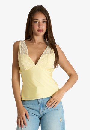 Donna con lunghi capelli castani che indossa una canotta gialla con bordi in pizzo e jeans azzurri strappati, in piedi con una mano sul fianco.