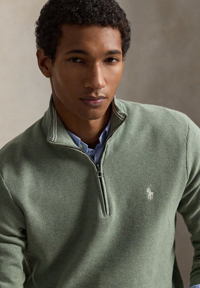 Roheline kudumiga poollukuga kampsun tekstuurse mustriga, millel on vasakul rinnal peen logo, kantud helesinise kraega särke peal.