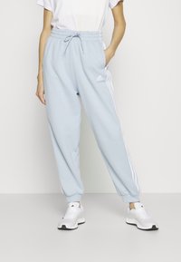 Pantalons de jogging bleu clair avec taille élastique, cordon de serrage, poches latérales et accents blancs à trois bandes. Le matériau semble doux et lisse.