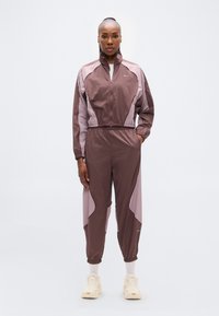 Καφέ και ροζ φόρμα με μπλοκ χρωμάτων, ψηλό γιακά, ελαστική μέση και στενές παντελόνι, με το λογότυπο της Nike στο στήθος.