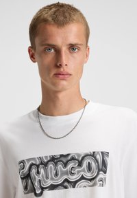 Hvid bomulds t-shirt med et stort sort og sølv abstrakt grafik centralt på brystet. Modellen bærer et sølvfarvet kædehalssmykke.