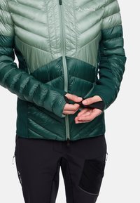 Giacca isolante verde con design a zig zag, dotata di una chiusura lampo frontale, polsini elastici e una consistenza piumosa. Abbinata a pantaloni neri.