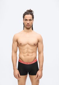 Μυώδης άνδρας με τρέσες που φοράει μαύρα μπόξερ Tommy Hilfiger, στέκεται μπροστά σε λευκό απλό φόντο.