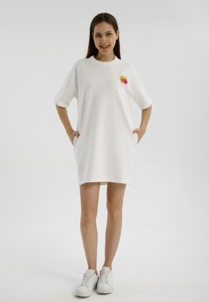 Jeune femme debout, les mains dans les poches, portant une robe t-shirt blanche oversize avec un motif coloré sur la poitrine et des baskets blanches.
