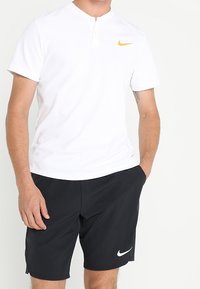 Camisa henley blanca con un logo de Nike amarillo, combinada con pantalones cortos negros que presentan un swoosh blanco de Nike. Ropa deportiva ligera y casual.