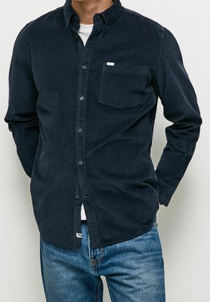 Mężczyzna ubrany w ciemnogranatową koszulę sztruksową na guziki, na białej koszulce i niebieskich dżinsach, stoi z rękami za plecami na jednolitym tle.