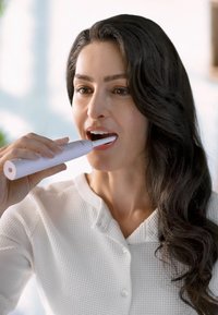 Spazzolino elettrico con un corpo elegante e bianco e una testina a punta, tenuto in una mano. L'utente sta spazzolando i denti, con lunghi capelli scuri visibili.