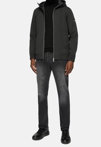 Men's zip-up jacket in dark gray fabric, featuring a hood, side pockets, and ribbed cuffs, paired with dark gray jeans and black sneakers.