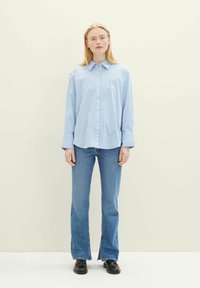 Camisa azul e branca com riscas, de botões e mangas longas, combinada com jeans claros de perna larga e sapatos pretos, em fundo neutro.