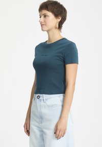 Türkises, eng anliegendes T-Shirt mit runder Halslinie und kurzen Ärmeln. Verfügt über ein kleines schwarzes Logo auf der Brust. Kombiniert mit hellblauen hoch taillierten Jeans.