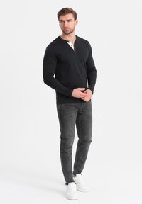 Zwart long-sleeve henley shirt met een contrasterende witte knoopsluiting en knopen, gecombineerd met vervaagde zwarte slim-fit jeans en witte sneakers.