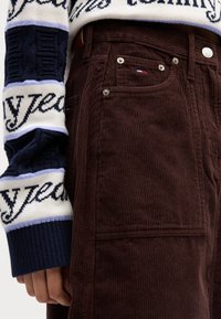 Pantaloni in corduroy marrone con righe verticali e rivetti metallici, abbinati a un maglione a maglia con motivo navy e crema.