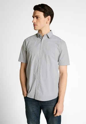 Mann trägt ein kurzärmliges hellgraues Hemd mit Knöpfen und dunkelblaue Jeans, steht mit einer Hand in der Tasche und schaut nach links.