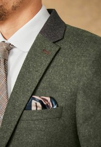 Blazer en laine verte avec un col sombre contrastant, orné d'un poche carré aux motifs bleus et roses, associé à une cravate à motifs.