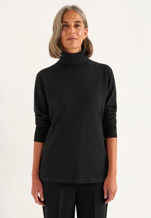 Vrouw met schouderlang grijs haar, die een zwarte coltrui en zwarte broek draagt tegen een effen lichte achtergrond.