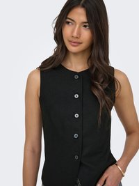 Femme aux longs cheveux ondulés porte un haut noir sans manches avec boutons, regardant sur le côté, la main posée sur la hanche, sur fond uni.
