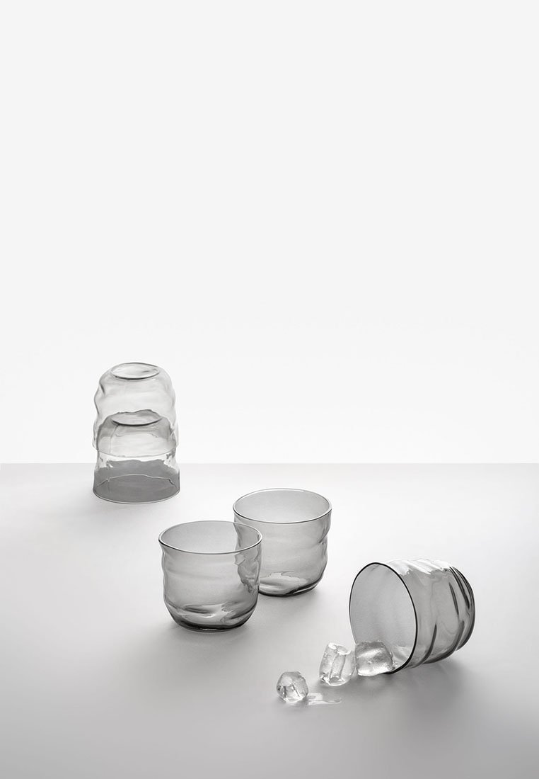 Cztery przezroczyste, teksturowane szklanki do picia na białej powierzchni, jedna przewrócona z kostkami lodu wypadającymi na zewnątrz.