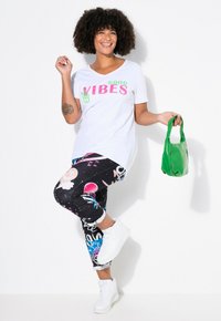 T-shirt blanche avec le texte « GOOD VIBES », leggings noirs avec des motifs graphiques colorés, sac à main vert et baskets montantes blanches.