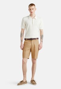 Polo di tessuto cremoso e strutturato, abbinato a pantaloni corti beige. Una cintura in pelle marrone e scarpe da barca marroni completano l'outfit.