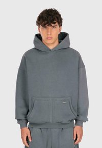 Junger Mann mit lockigem Haar, der einen lockeren grauen Kapuzenpullover mit Fronttaschen und passende graue Hose trägt, steht vor einem einfarbigen hellen Hintergrund.