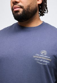 Κοντινό πλάνο ενός άνδρα με γένια που φοράει μπλε πουκάμισο με μικρό λευκό και κίτρινο κείμενο στο στήθος.