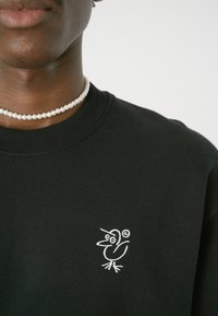Schwarzer Sweatshirt mit einem kleinen, weißen, gestickten Vogeldesign auf der linken Brust. Das Material wirkt weich und strukturiert mit einem Rundhalsausschnitt.