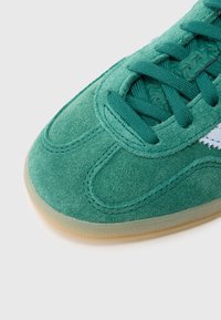 Ténis de camurça verde com biqueira arredondada, parte superior texturizada e atacadores a condizer. Apresenta uma sola de goma translúcida e detalhes subtis de costura.