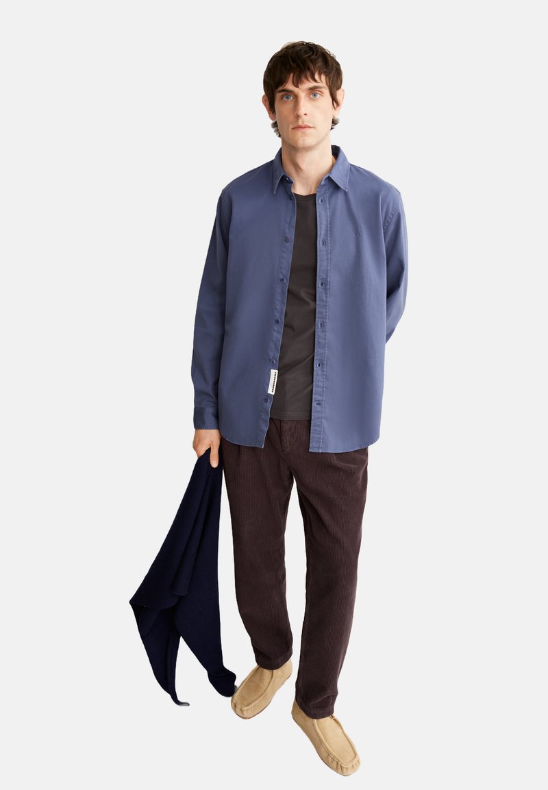 Een lichtblauwe overhemden van katoen, gecombineerd met een donkergrijze T-shirt, een paar bruine corduroyen broek en tan kleurige moccasin schoenen, met een marineblauwe trui in de hand.