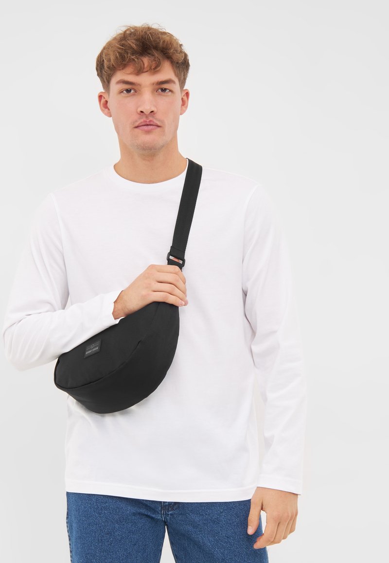 Borsa a tracolla nera realizzata in tessuto resistente, forma arrotondata, con una tracolla e un'etichetta frontale. Indossata sopra una maglietta bianca a maniche lunghe.